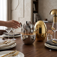 A polished silver bowl-shaped utensil holder filled with metallic cutlery sits on a wooden dining table, alongside a large reflective gold egg-shaped container and place settings. A person's hand is visible reaching toward the utensils, and the scene is set in a real interior dining environment.