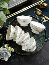 A set of white porcelain tea or dessert bowls with delicate floral illustrations and gold trim, arranged on a dark green marbled tray with gold-rimmed edges. The tray is styled on a dark marble surface with decorative props including a gold spoon, a gold teapot, and green leaves.