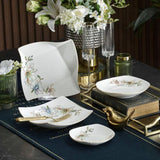 A set of white ceramic tableware with delicate floral and bird illustrations, styled on a dark table runner with gold accents, books, and glassware.