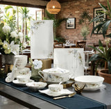 A coordinated ceramic tableware set with floral prints is arranged on a dining table in a restaurant setting. The set includes plates, bowls, cups, saucers, napkins, and a tureen, all in white with delicate blue and green floral detailing.
