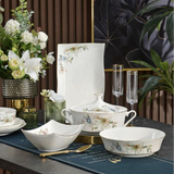 A set of white porcelain dinnerware with delicate blue and green floral patterns, arranged on a dark table runner with gold accents. The set includes a square platter, a lidded soup bowl, a lidded casserole dish, a bowl with a handle, a shallow square bowl, and two clear champagne flutes, styled with a gold knife and surrounded by floral arrangements and green plants.