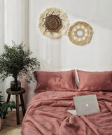 A bedroom scene showing a bed made with dusty rose pink bedding, including a duvet cover and matching pillowcases. The top panel includes wall decor and a laptop on the bed, while the bottom panel shows a wider view of the styled room with plants and a window.