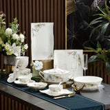 An elegant porcelain dinnerware set with floral gold and blue-green illustrations, displayed on a dark table runner with gold accents, glassware, and decorative flowers against a dark wood slatted background.