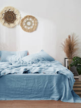 Two bedroom scenes showing bedding in different colorways: a top view with light blue sheets and pillows, and a bottom view with gray sheets and pillows, both styled with wall decor, pampas grass, and plants.