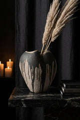 A dark ceramic vase with a textured matte black finish and beige dripped patterns, holding dried pampas grass, styled on a black marble surface with books and lit candles in the background.