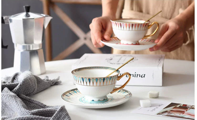 Tea cups with spoons on saucers, a silver Moka pot, and a person holding a cup on a table.