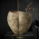 A large textured ceramic vase with a ribbed, basket-like pattern in beige and brown tones, displayed on a dark stone pedestal against a dark background with dried lotus stems in the opening.