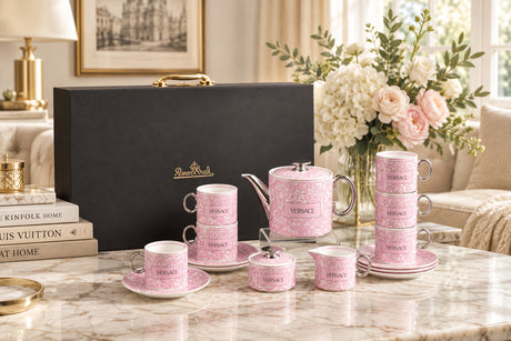 A pink Versace teacup and teapot set arranged on a marble table in a styled interior, with a large black presentation box behind it. The cups, saucers, teapot, and small containers are decorated with a light pink damask pattern and the Versace logo, while the background includes flowers, a lamp, framed art, and a sofa.
