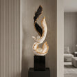 A decorative sculptural bird figure on a black pedestal, shown in an interior setting with wood slat walls and a window in the background. The sculpture is primarily white and black with metallic gold feather accents and clear crystal-like spheres.