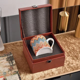 Colorful zigzag-patterned mug in a brown leather box on a wooden surface.