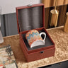 Colorful zigzag-patterned mug in a brown leather box on a wooden surface.