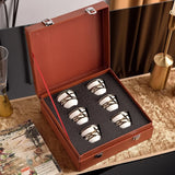Set of four silver and white ceramic cups in a brown leather case on a decorative surface.