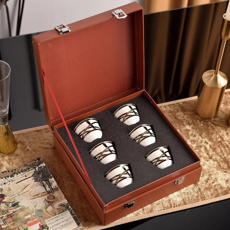 Set of four silver and white ceramic cups in a brown leather case on a decorative surface.