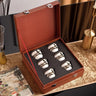 Set of four silver and white ceramic cups in a brown leather case on a decorative surface.