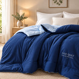A blue silk quilt comforter set on a bed with white pillows and a light blue pillowcase, styled in a minimalist bedroom setting with a small plant on a side table.