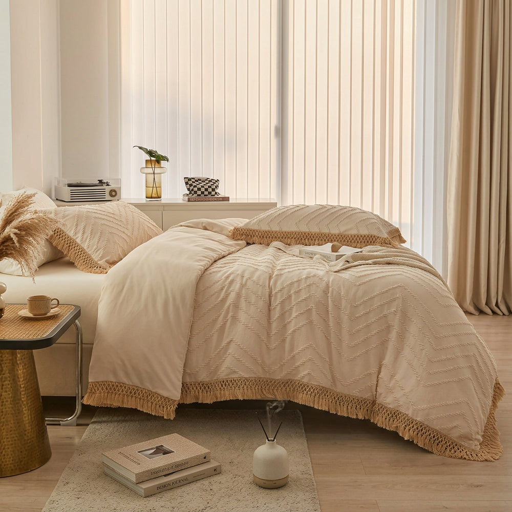 A neatly made bed in a bright bedroom setting, styled with a cream and beige chevron-patterned duvet cover, matching pillowcases, and textured beige fringed borders. The room features white vertical blinds, a small side table with a record player and vase, and a light wood floor.