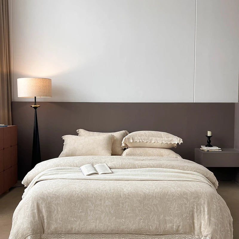 A neatly made bed in a modern bedroom, dressed in light beige patterned bedding with matching pillows and a folded throw blanket. The headboard is dark brown, with a floor lamp on the left and a bedside table on the right.