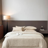 A neatly made bed in a modern bedroom, dressed in light beige patterned bedding with matching pillows and a folded throw blanket. The headboard is dark brown, with a floor lamp on the left and a bedside table on the right.