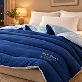 A navy blue silk quilted comforter with white piping and a white printed logo, styled on a bed with cream pillows and a hand lifting the corner to show its soft texture.