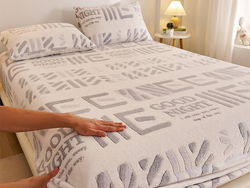 Bed with a textured blanket featuring text, hands touching the blanket, and a nightstand in the background.