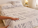Bed with a textured blanket featuring text, hands touching the blanket, and a nightstand in the background.