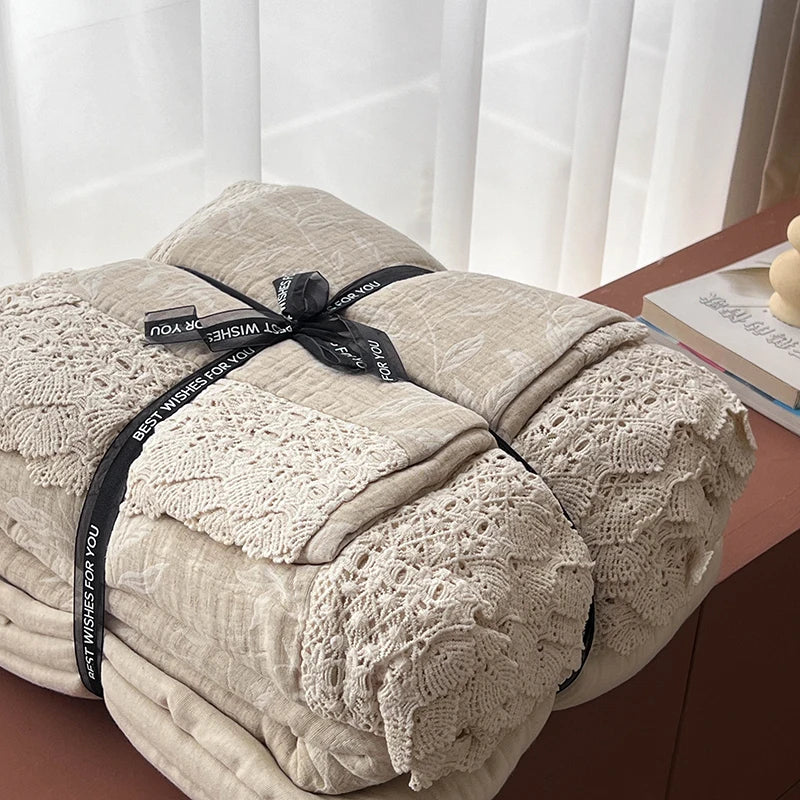 A folded beige quilted blanket with a matching lace-trimmed pillow, wrapped in a black ribbon printed with 'BEST WISHES FOR YOU', displayed on a wooden table next to a book.