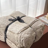 A folded beige quilted blanket with a matching lace-trimmed pillow, wrapped in a black ribbon printed with 'BEST WISHES FOR YOU', displayed on a wooden table next to a book.