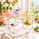 Four elegant clear coupe glasses filled with colorful cocktails arranged on a marble table in a bright indoor setting. The drinks are pink, lavender, pale yellow, and light green, garnished with citrus slices, berries, lavender sprigs, and mint leaves, with flowers and lemons in the background.