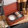 Set of colorful ceramic cups and saucer in a brown leather case on a decorative surface.