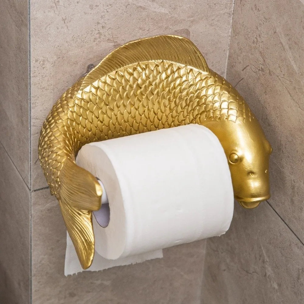 A gold fish-shaped toilet paper holder mounted on a beige tiled wall, holding a white roll of toilet paper.