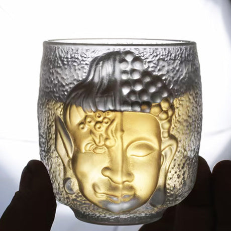Glass tumbler with Buddha face design held by a hand against a white background