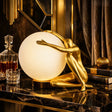 A decorative table lamp featuring a gold-finished sculptural figure kneeling behind a large glowing white globe shade, placed on a dark marble surface in an ornate interior setting.