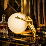 A decorative table lamp featuring a gold-finished sculptural figure kneeling behind a large glowing white globe shade, placed on a dark marble surface in an ornate interior setting.