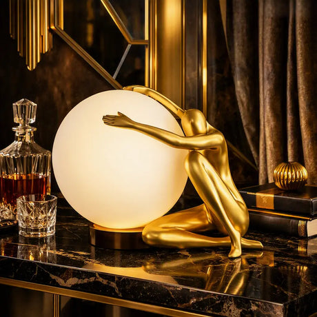 A decorative table lamp featuring a gold-finished sculptural figure kneeling behind a large glowing white globe shade, placed on a dark marble surface in an ornate interior setting.