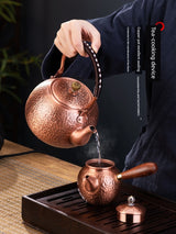 Copper teapot being used to pour tea into another copper teapot on a wooden tray.