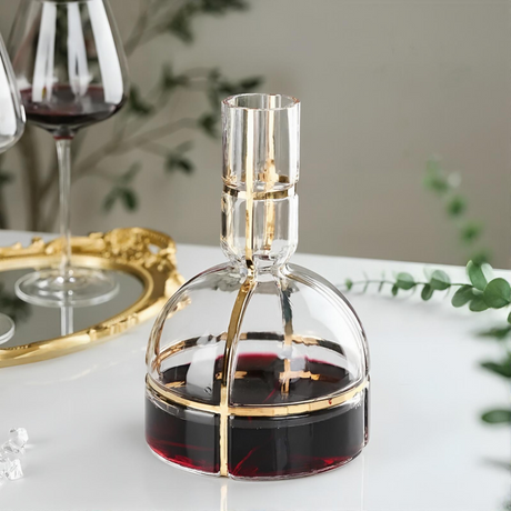 Clear wine decanter with red wine on a white surface, with wine glasses and a decorative tray in the background.