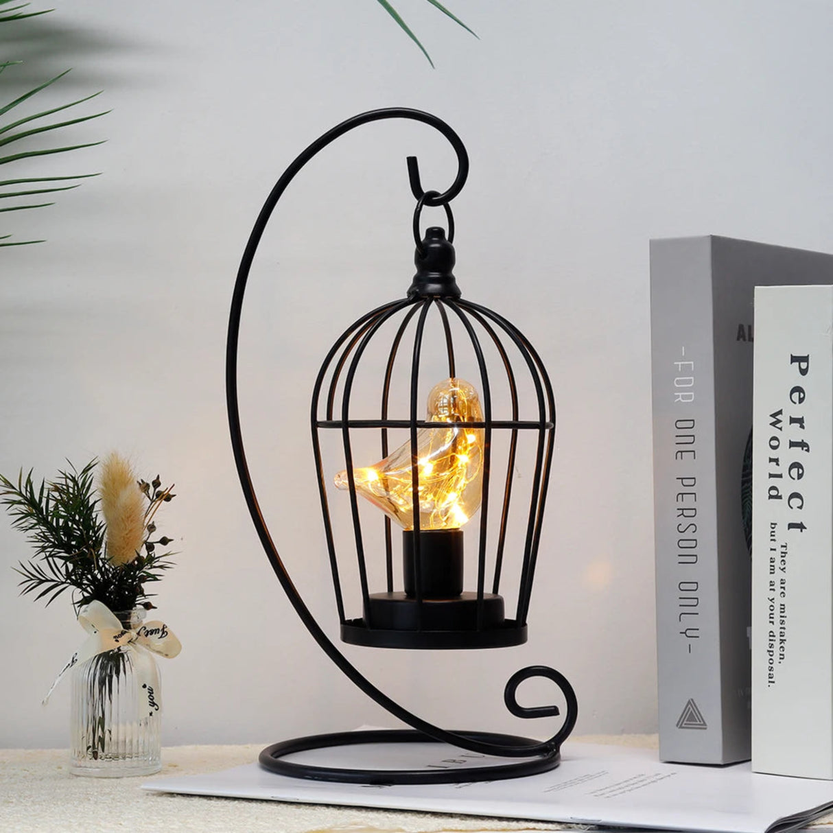 Decorative black metal cage with a light bulb inside, placed on a surface with a plant and book in the background.