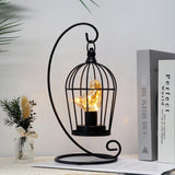 Decorative black metal cage with a light bulb inside, placed on a surface with a plant and book in the background.