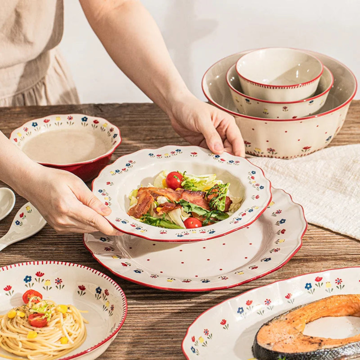 Person holding a bowl of salad with decorative ceramic plates on a wooden table.